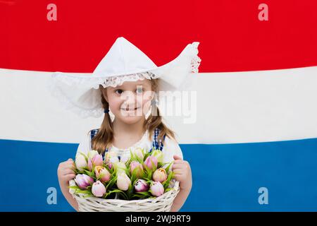 Bambina olandese che indossa il tradizionale costume nazionale, l'abito e il cappello che reggono un cestino di tulipani sulla bandiera dei Paesi Bassi. Bambino con souvenir Foto Stock