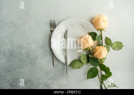 Vista dall'alto di un elegante tavolo con piatto bianco, posate in argento e delicate rose alla luce del sole. Foto Stock