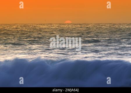 Una tranquilla scena oceanica al tramonto con colori vivaci e dolci onde il sole si tuffa sotto l'orizzonte, dipingendo il cielo con sfumature di arancione Foto Stock