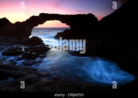 Un'alba serena irrompe su El Hierro, proiettando una luce soffusa attraverso un arco di roccia vulcanica naturale sopra le vorticose acque marine Foto Stock