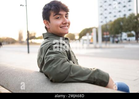 Ritratto di un brunetto brunetto con giacca e jeans seduti sulla panchina contro un ambiente urbano sfocato che guarda la fotocamera Foto Stock