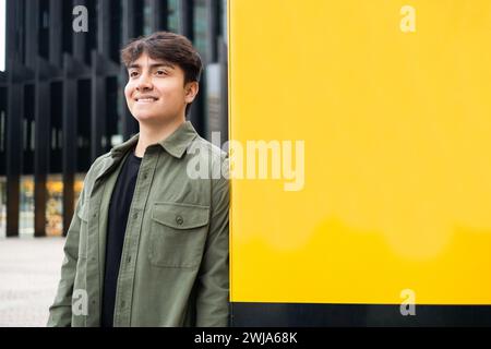 Uomo felice in abiti informali che si trovano vicino a un moderno edificio dalle pareti gialle e che guardano lontano in città Foto Stock