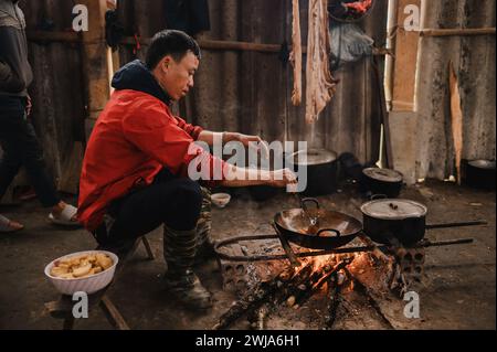 Vista laterale di un ragazzo in abiti informali seduto sulla panchina e che mescola il cibo con la spatola nella vecchia cucina malandata del Vietnam Foto Stock