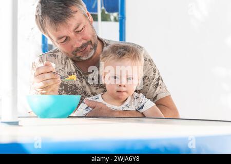 Un padre amorevole sta nutrendo il suo bambino con la sindrome di Down da una ciotola blu a un tavolo all'aperto, condividendo un tenero momento in famiglia Foto Stock