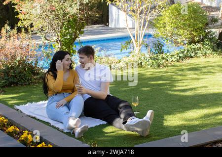 Giovane uomo e donna felici in abiti casual con sorriso sorridente seduti sul prato vicino alla piscina mentre si guardano l'un l'altro Foto Stock