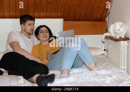 Corpo pieno di allegri giovani uomini e donne in abiti casual sdraiati sul letto mentre usano il laptop per navigare in camera da letto Foto Stock