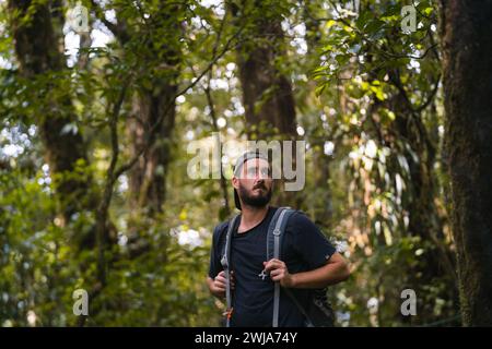 Un escursionista maschile barbuto con uno zaino che guarda lontano in mezzo a una fitta vegetazione verde della foresta, mostrando un senso di avventura e scoperta Foto Stock