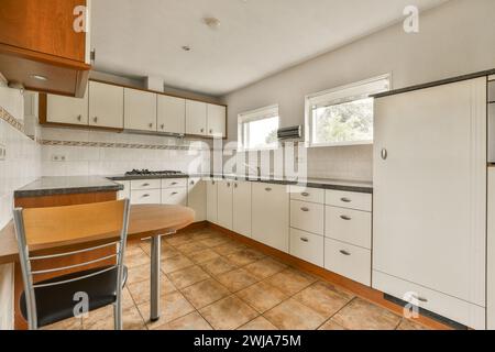Una ben illuminata e spaziosa cucina con armadi in legno bianco, pavimento piastrellato marrone e una piccola area da pranzo con tavolo e sedia. Foto Stock