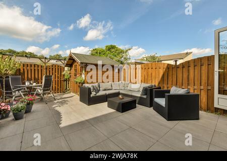 Un patio moderno con eleganti mobili da giardino, capannone in legno e piante lussureggianti sotto un cielo azzurro e soleggiato. Foto Stock