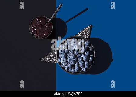 Vista dall'alto dei mirtilli in una ciotola con marmellata di mirtilli su un tovagliolo geometrico, su uno sfondo blu e nero che crea un effetto d'ombra dinamico Foto Stock