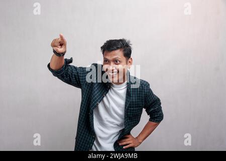 Un uomo asiatico con una camicia a scacchi verde si erge e punta il dito destro sullo sfondo isolato "guardando te" Foto Stock