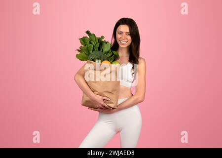 Una donna sorridente in un abito sportivo bianco tiene con sicurezza una borsa della spesa piena di frutta e verdura verdi Foto Stock