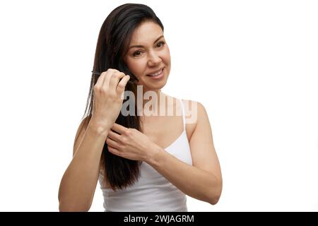 Ritratto di una donna di mezza età che pettina i suoi capelli lunghi sani e setosi guardando lontano sullo sfondo bianco dello studio. Foto Stock
