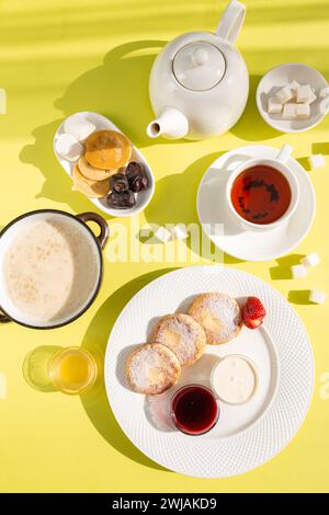 Assapora una deliziosa colazione con frittelle di formaggio, farinata d'avena cremosa, tè aromatico, miele dorato, marmellata, e frutta fresca su un tavolo giallo soleggiato, c Foto Stock