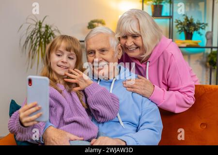 Nonno caucasico, nonna e piccola nipote che fanno videochiamate online su smartphone a casa. Ragazza con i nonni anziani che parla con un'app chat tramite webcam Internet seduta sul divano della camera. Foto Stock