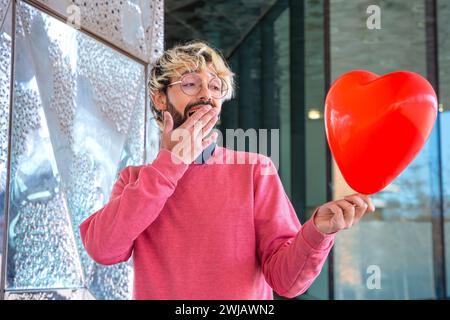 Un giovane sorpreso in piedi reggendo un palloncino a forma di cuore. Amore Foto Stock