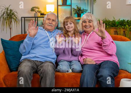 La coppia anziana dei nonni con la nipotina, le onde dei bambini salutano, un saluto rilassante sul divano nel soggiorno di casa. Nipote e famiglia che fanno vlog trascorrendo il tempo libero insieme in appartamento. Foto Stock