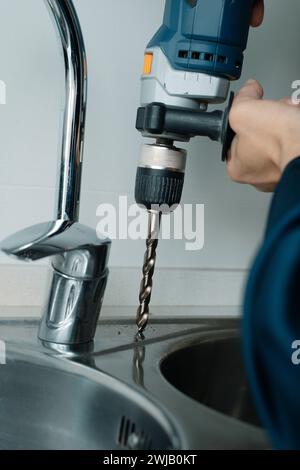 primo piano di un uomo caucasico, indossa tute blu, usando un trapano per fare un foro in un lavandino da cucina in metallo Foto Stock