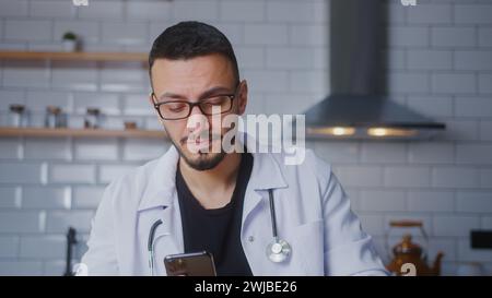 Il medico maschile indossa l'uniforme bianca con lo stetoscopio che naviga su Internet sul telefono cellulare, controlla la posta elettronica, legge notizie sui media, scorrendo i social network, utilizzando Foto Stock