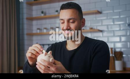 Un uomo sorridente siede nella cucina moderna di casa, tenendo un salvadanaio e mette dentro la moneta Foto Stock