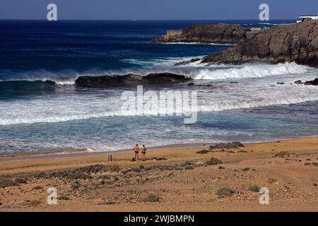 Due uomini in piedi a parlare su una piccola spiaggia sabbiosa con mare selvaggio e onde atlantiche che si infrangono, El Cotillo, Fuerteventura, presa nel novembre 2023 Foto Stock