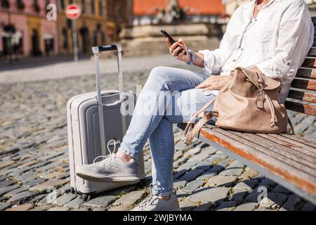 La donna che viaggia con i bagagli è seduta e riposa sulla panchina in città. Turista femminile che utilizza uno smartphone Foto Stock