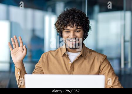 Foto ravvicinata di un giovane indiano che parla tramite collegamento video. È seduto in ufficio a una scrivania con una cuffia e davanti a un computer portatile. Sorridendo e sventolando davanti alla telecamera. Foto Stock