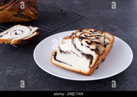 Pezzi di panino intrecciato fresco fatto in casa con semi di papavero su piatto bianco, sfondo nero Foto Stock