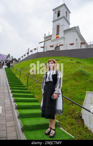 Giovane donna in costume tradizionale popolare di fronte a una chiesa su una collina verde, donna in costume tradizionale, Vadu Motilor, Alba, Transilvania, Romania Foto Stock