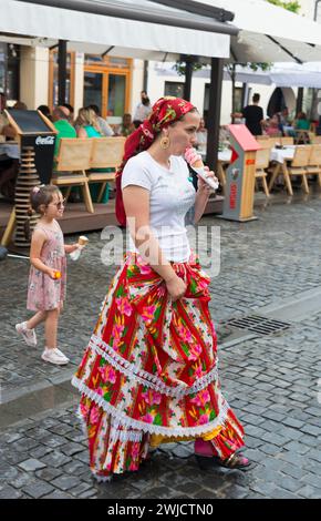 Donna in abito tradizionale che mangia gelato in una strada trafficata, bambino sullo sfondo, giovane donna in abito tradizionale, Sibiu, Transilvania, Romania Foto Stock