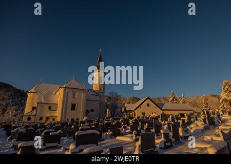 Chiesa di santa maria nel villaggio di Ljubno ob Savinji nelle prime ore del mattino durante la metà dell'inverno, piena di neve, tombe e chiesa ricoperta di sostanziale consistenza Foto Stock