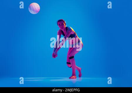 Giocatrice di Beach volley che si prepara a servire la palla su sfondo blu sfumato con luce al neon rosa. Foto Stock