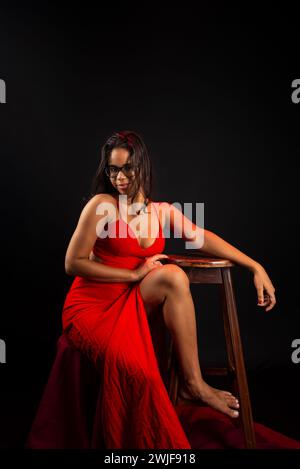 Salvador, bahia, Brasile - 09 dicembre 2023: Ritratto di una bella giovane donna che indossa gli occhiali e in abito rosso seduta in posa per una foto. Isolato su Foto Stock