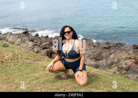 Salvador, bahia, Brasile - 06 gennaio 2024: Bella donna dai capelli ricci che indossa occhiali da sole accovacciati su una collina contro il mare e le rocce nel backgrou Foto Stock