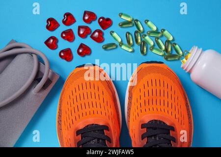 Tonometro per la misurazione della pressione sanguigna, scarpe sportive arancioni, capsule gialle di vitamina D e cuori di vetro rosso sparsi su sfondo blu. Vitamina Foto Stock