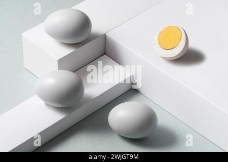 Uova bianche e un uovo tagliato a metà su sfondo grigio a strati con piedistalli, STILL Life, scatto ad angolo alto Foto Stock