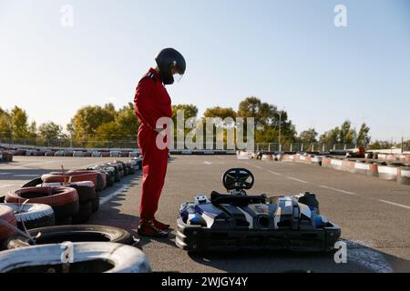 Autista che indossa tute da corsa e casco in piedi nelle vicinanze dell'auto da corsa di kart Foto Stock