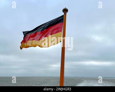 Una bandiera tedesca che sventola nel Mare del Nord, tra il mare turbolento e il cielo nuvoloso in una fredda giornata invernale a bordo del traghetto diretto all'isola di Langeoog Foto Stock