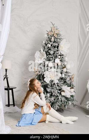 Una giovane donna siede sul pavimento in una stanza accogliente, ammirando un albero di Natale splendidamente decorato. L'albero è decorato con ornamenti bianchi e dorati Foto Stock