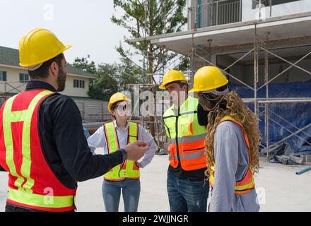 Un gruppo di operai edili che indossa un elmetto e un giubbotto di sicurezza si riunisce sui piani di un cantiere con attrezzi e attrezzature intorno a loro. Foto Stock