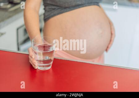 Abbracciando i benefici vitali dell'acqua durante la gravidanza, una donna incinta sta in cucina con un bicchiere, evidenziando il ruolo cruciale dell'idratazione in Foto Stock