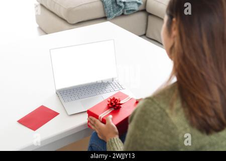 Giovane donna birazziale a casa, con un regalo con un portatile sul tavolo, durante una videochiamata Foto Stock
