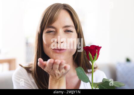 Una donna caucasica di mezza età sta scherzosamente soffiando un bacio mentre tiene in mano una rosa rossa Foto Stock