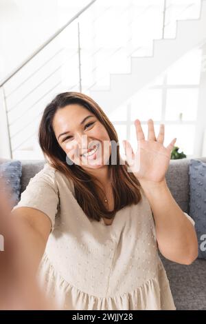 Una giovane donna birazziale ondeggia alla telecamera in un ambiente domestico luminoso durante una videochiamata Foto Stock