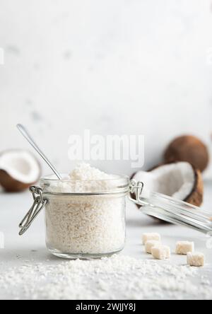 Vaso in vetro con scaglie di cocco dolci grattugiate e noci di cocco maturo su un tavolo leggero. Foto Stock