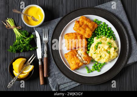 filetto di pesce fritto croccante con purè di patate e piselli verdi sul piatto su un tavolo di legno nero con posate rustiche, senape, limone, vista orizzontale da Foto Stock