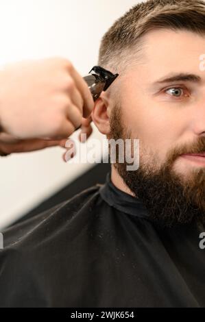 Il barbiere rasa il contorno della linea ovale con un regolacapelli sulla testa del cliente. Un uomo con la barba si fa tagliare i capelli su una sedia da barbiere. Foto Stock