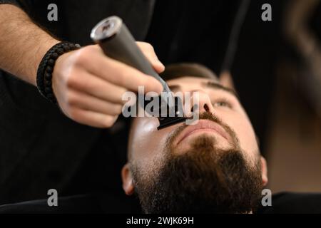 Un barbiere rifinisce i baffi di un cliente caucasico con un trimmer. Foto Stock