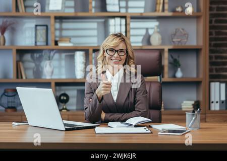 Donna matura e allegra in abito da lavoro che dà un gesto con il pollice mentre lavora alla sua scrivania con laptop e smartphone. Foto Stock