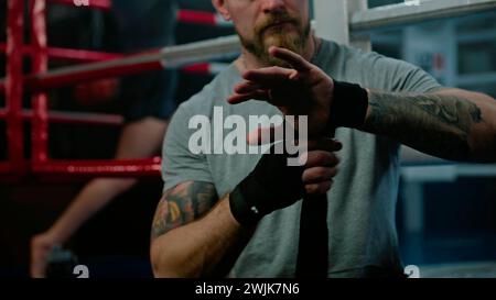 Uomo adulto seduto vicino all'anello di pugilato e avvolge le mani con una benda per le arti marziali L'atleta si prepara a combattere, gareggiare o allenarsi in box gym. Boxer esegue la boxe ombra sullo sfondo. Primo piano. Foto Stock